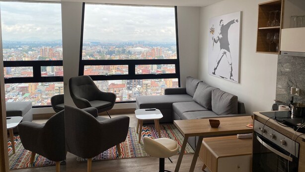 A modern living room with a city view, a grey sofa, a small table, and a kitchen area with a stove.