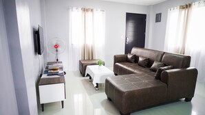 A modern living room with a brown leather sofa, a white side table with books, a standing fan, and a small plant on a white table. - Cozy and homelike accommodations (Olongapo City)
