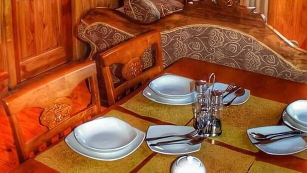 Een houten eetkamer met een grote tafel gedekt voor vier personen, een beklede bank en een raam met gordijnen.