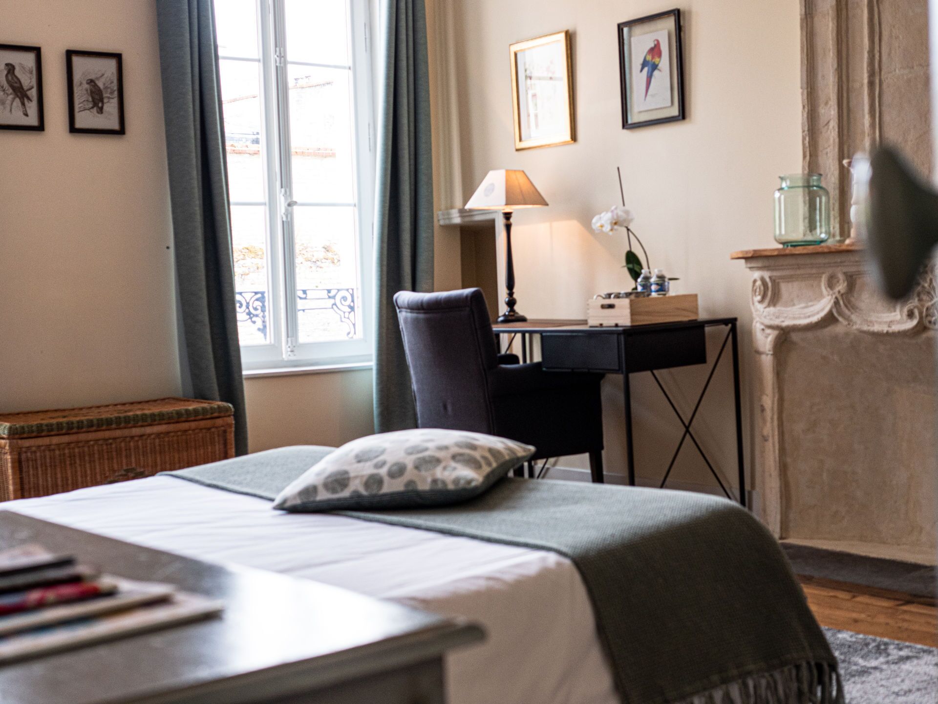 Un dormitorio ordenado con cama, escritorio y silla. Hay cuadros enmarcados en la pared, una lámpara en el escritorio y una ventana con cortinas.