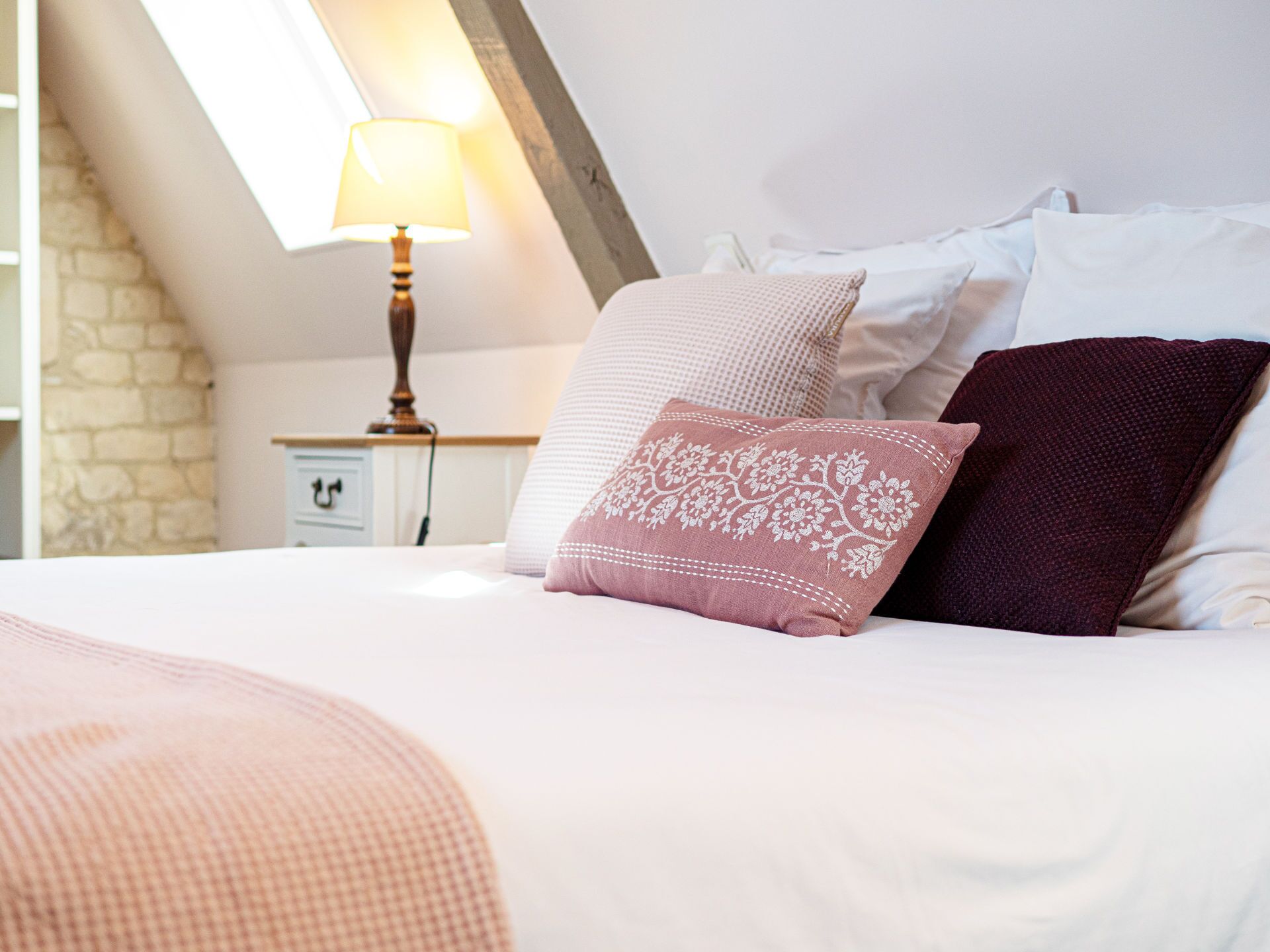 Una cama bien hecha con un cojín estampado en rosa y blanco, un cojín rojo oscuro y un cojín blanco. Hay una lámpara en una mesita de noche, con una pared de piedra al fondo.