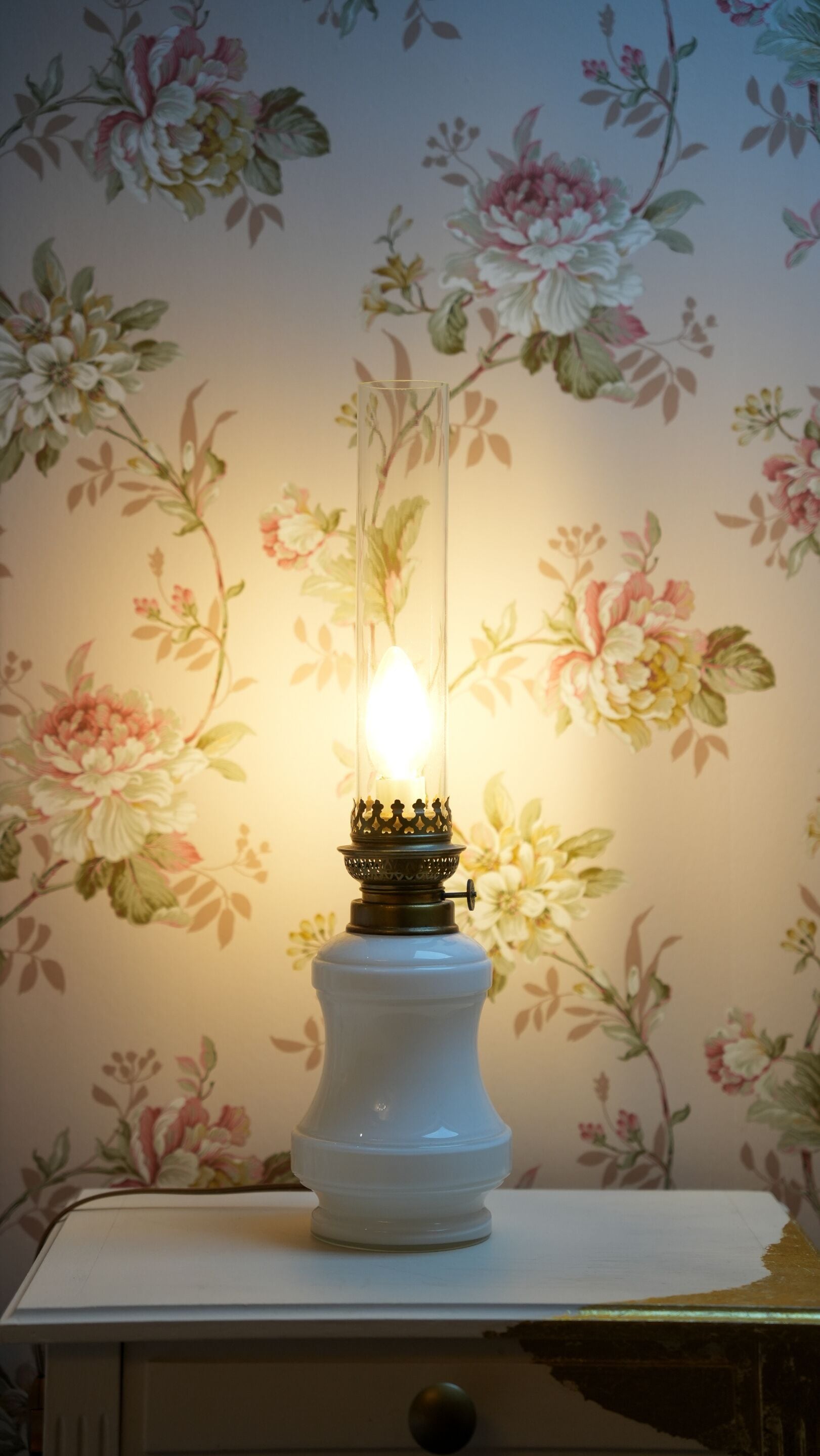 A white table lamp with a clear glass shade and a lit candle inside, placed on a white table against a floral wallpaper background.