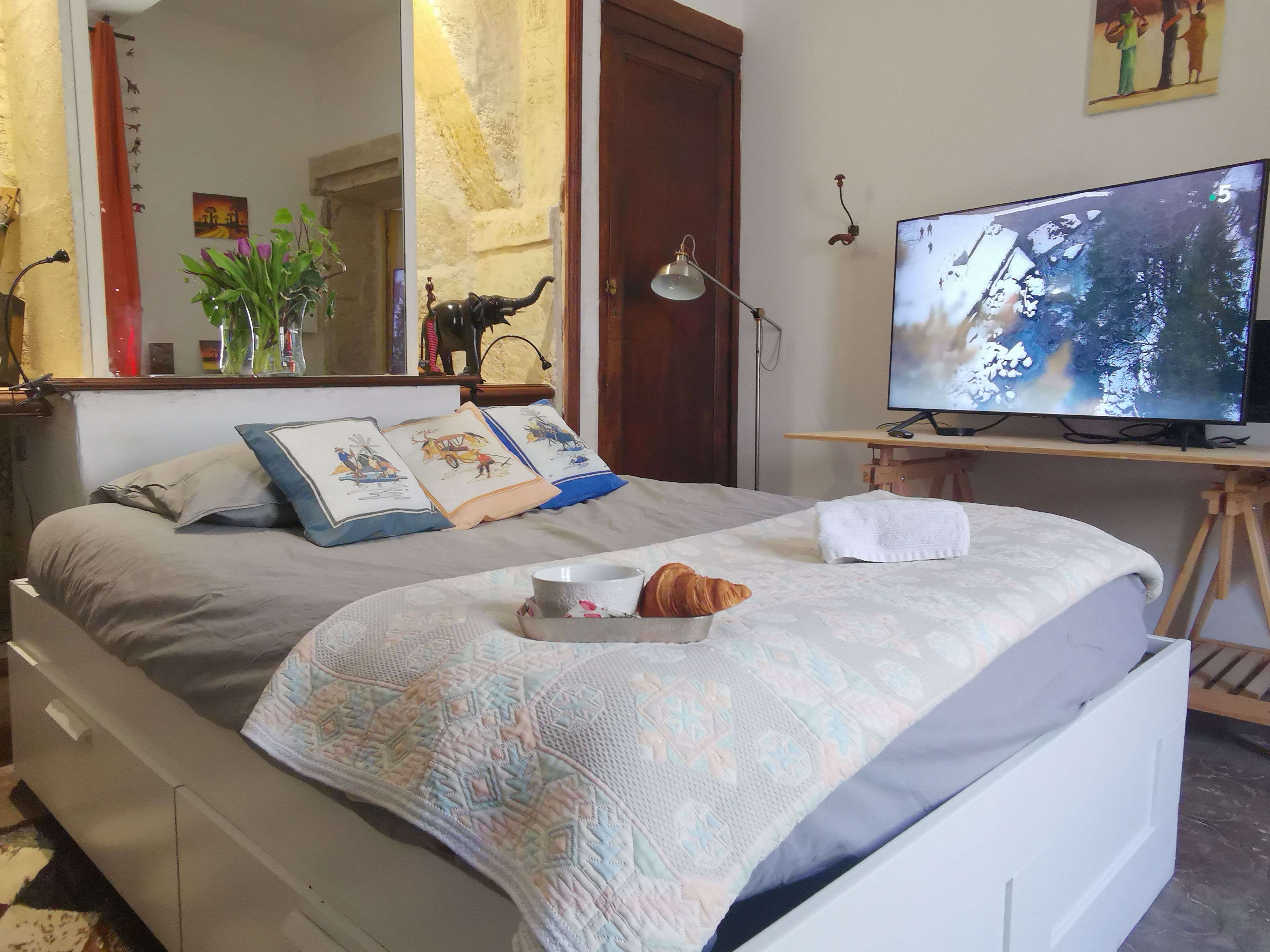 Ein ordentlich bezogenes Bett mit einer gemusterten Decke, ein Nachttisch mit einer Lampe, ein Fernseher und ein kleiner Tisch mit einem Croissant und einer Tasse.