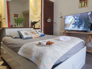 Ein ordentlich bezogenes Bett mit einer gemusterten Decke, ein Nachttisch mit einer Lampe, ein Fernseher und ein kleiner Tisch mit einem Croissant und einer Tasse.