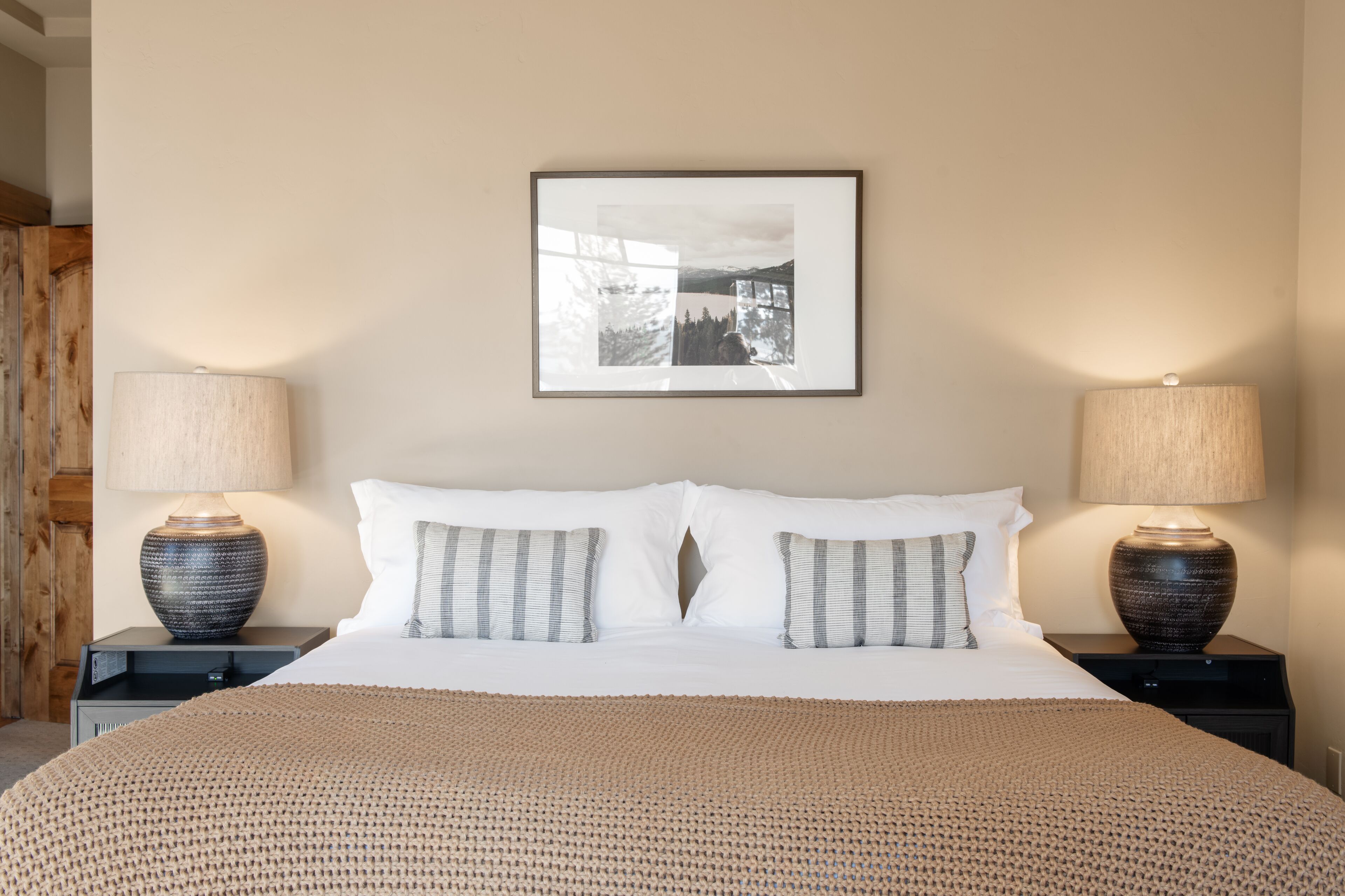 Un letto rifatto con cuscini bianchi e a righe, due abat-jour e un quadro incorniciato appeso al muro.