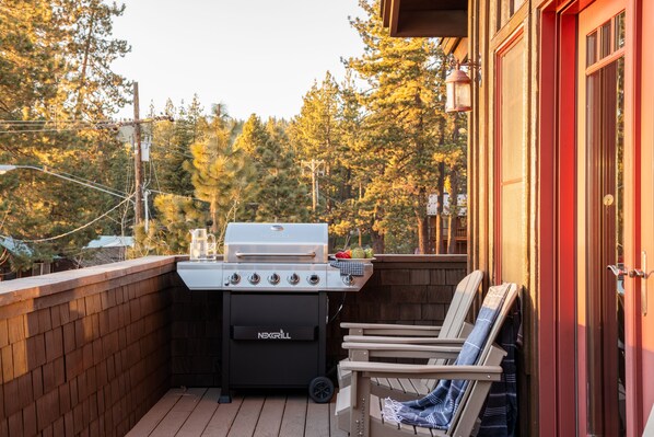 A barbecue grill on a wooden deck with a brick wall, a wooden chair, and a red door.