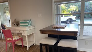 A dining area with a wooden table and two stools, a white desk with a chair, and a window with a view of a parked car and greenery outside. - Flip Flop 1 (Sanibel)
