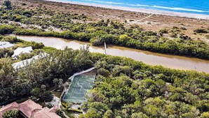 Luftaufnahme eines Küsteneigentums mit Tennisplatz, dichter Vegetation und Strand im Hintergrund.