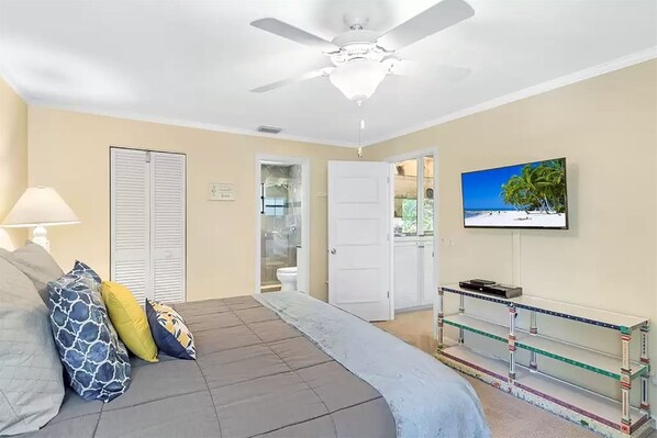 A bedroom with a bed, a TV, a ceiling fan, a bathroom door, and a glass table with a TV on it.