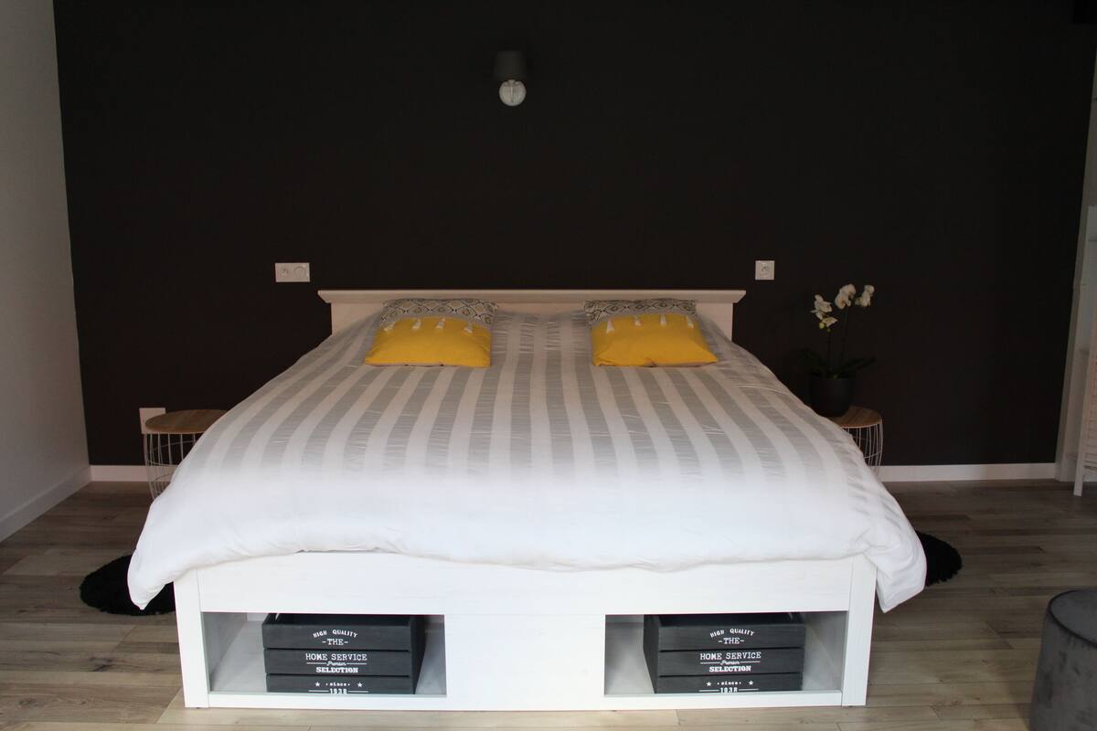 Een bed met een wit hoofdeinde en twee gele kussens, met aan weerszijden twee kleine nachtkastjes met boeken.