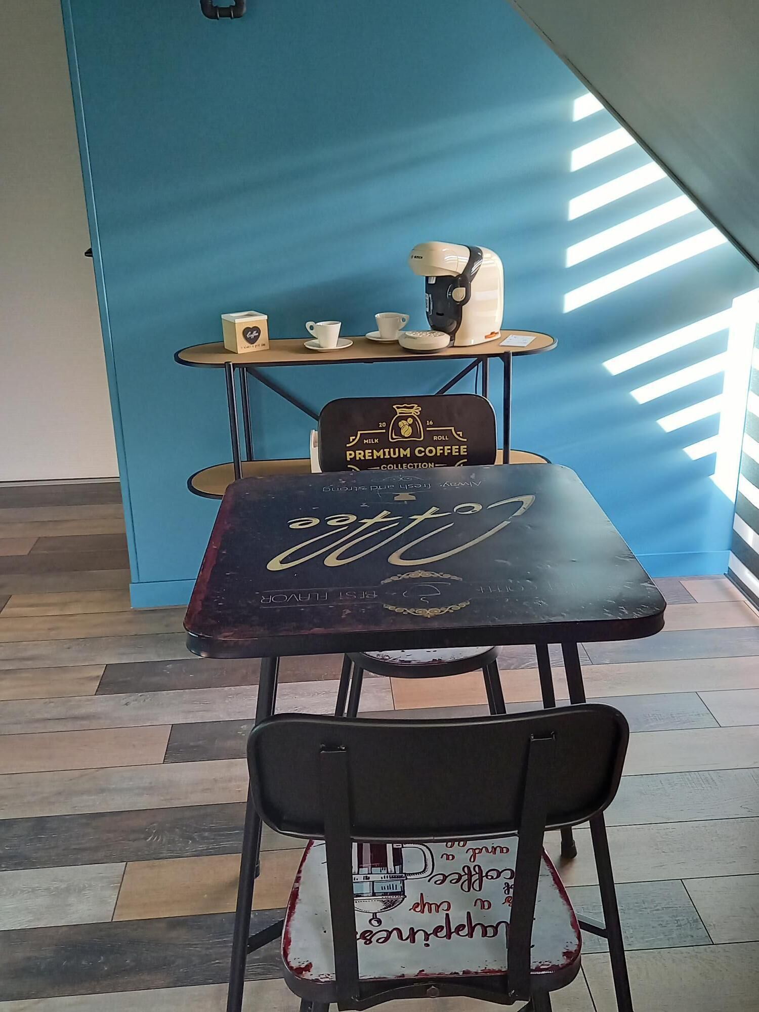 A table with a coffee machine and cups, a chair with a coffee-themed design, and a blue wall with a geometric pattern.