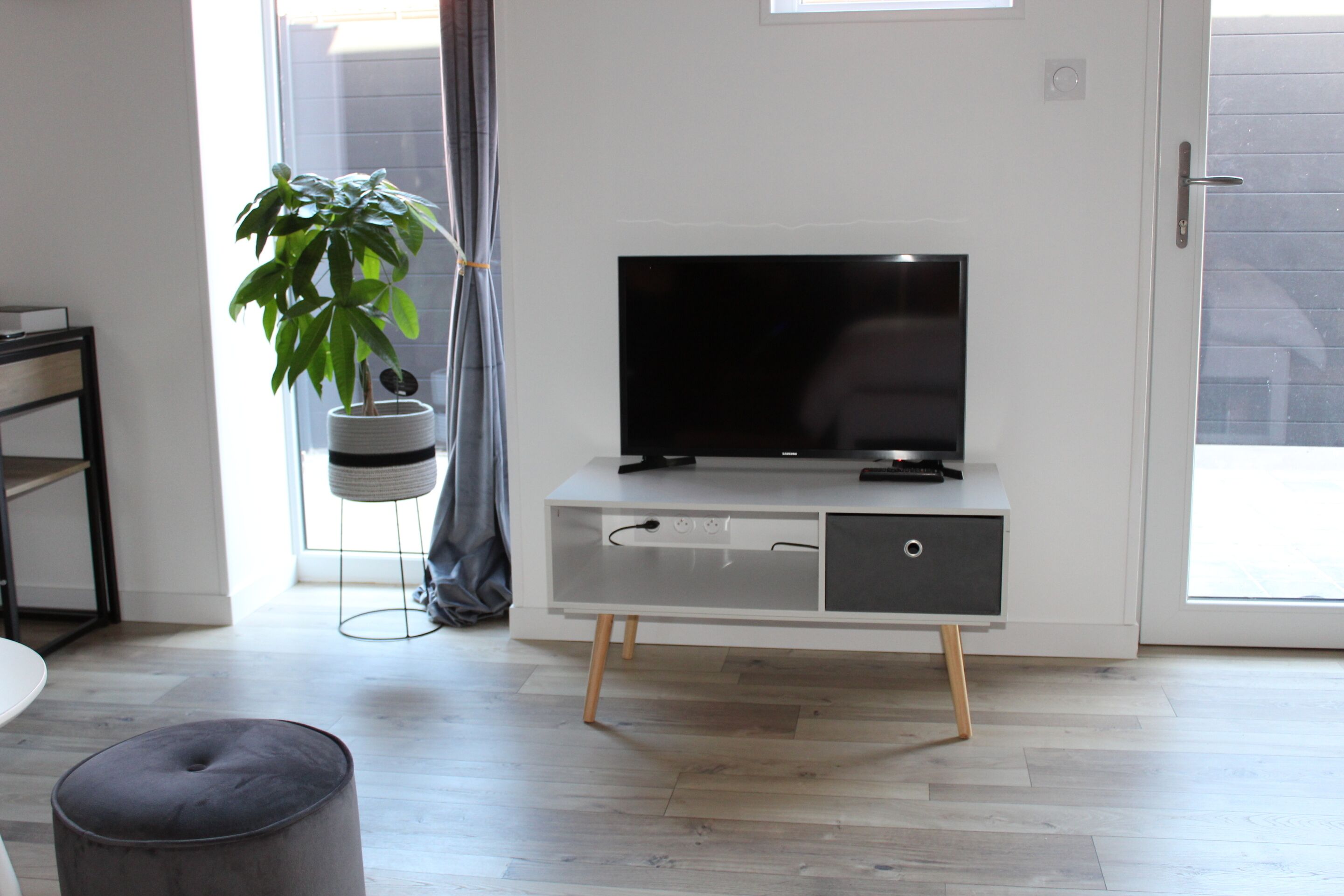 Een moderne woonkamer met een flatscreen televisie op een wit kastje met houten poten, een grijze poef en een potplant.