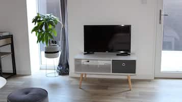 Een moderne woonkamer met een flatscreen televisie op een wit kastje met houten poten, een grijze poef en een potplant.