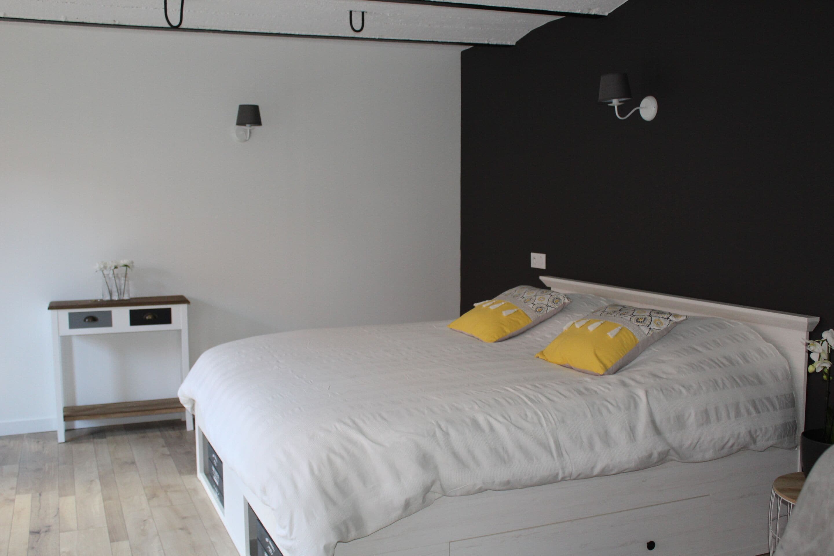 A bedroom with a white bed, two yellow pillows, a small bedside table, and a wall-mounted lamp.