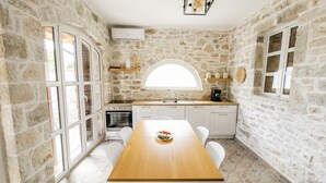 A kitchen with stone walls, a wooden table, white chairs, a large window, and a built-in oven. - Vila Leon - Old Qeparo (Upper Qeparo)