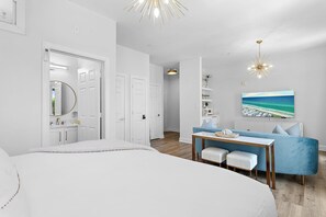 A modern bedroom with a large bed, a desk with a chair, a blue sofa, and a painting of a beach scene.