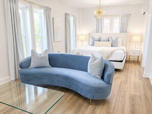 Et moderne soveværelse med en buet blå sofa, en hvid seng med mange puder, et glascaffe bord og en lysekrone.