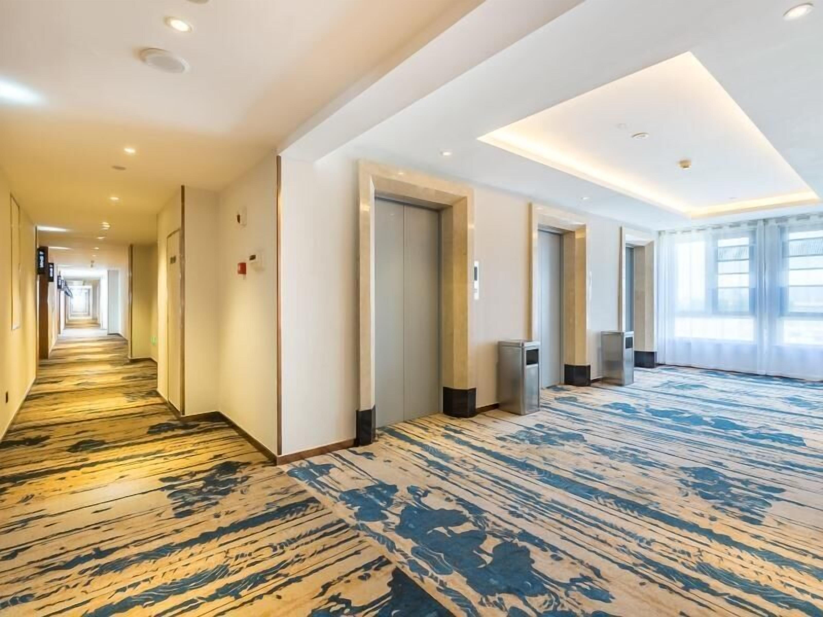 A hotel corridor with a patterned carpet, recessed lighting, and doors leading to rooms.