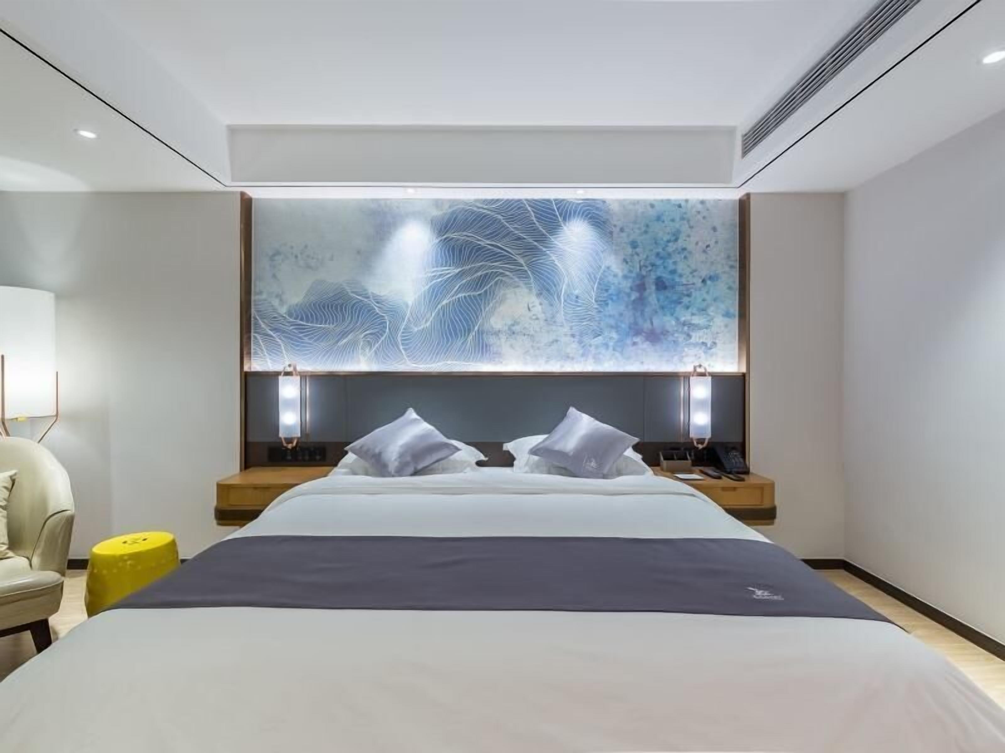 A modern hotel room with a large bed, two bedside tables, a wall art piece, and a chair.