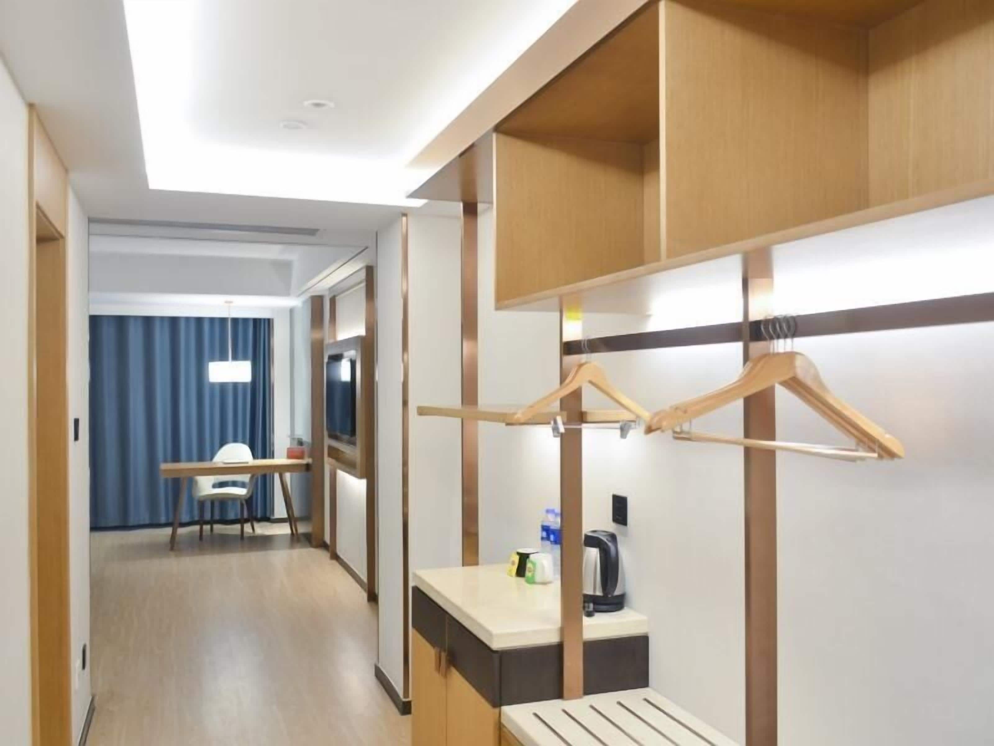 A hotel room with a wooden wardrobe, a desk with a chair, a bed, and a bedside table with a lamp.