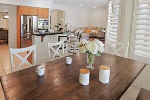 A modern kitchen with stainless steel appliances, a dining area with a wooden table and white chairs, and a living room with a sofa and decorative flowers.