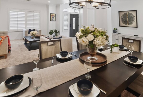 A modern dining room with a long table set for six, a vase of flowers, and a chandelier.