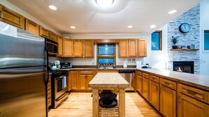A modern kitchen with wooden cabinets, a stainless steel refrigerator, a wooden table, and a stone accent wall. - 2027 OPEN! (3 Nt Min) Harbert Home With Private Hot Tub Steps To Beach! (Harbert)