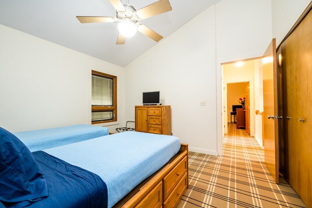A bedroom with two beds, a ceiling fan, a TV on a wooden cabinet, and a hallway leading to a bathroom.