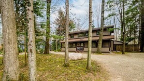A modern two-story house surrounded by tall trees and a gravel driveway. - 2027 OPEN! (3 Nt Min) Harbert Home With Private Hot Tub Steps To Beach! (Harbert)