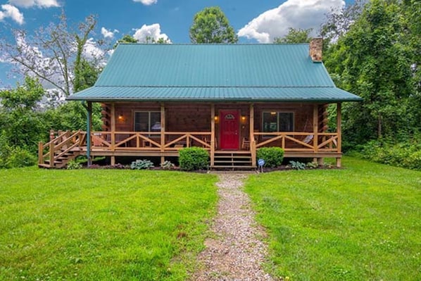 A log cabin with a green metal roof, wooden porch, and a gravel path leading to the front door.