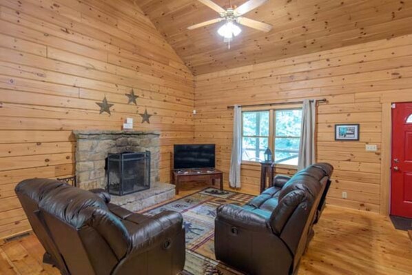 A log cabin interior with a stone fireplace, leather sofas, a television, and a ceiling fan.