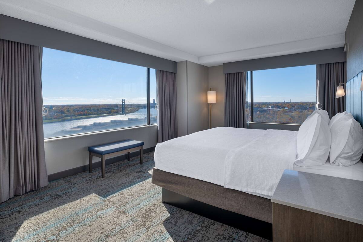 Een hotelkamer met een groot bed, een bankje en uitzicht op een stadslandschap en rivier.