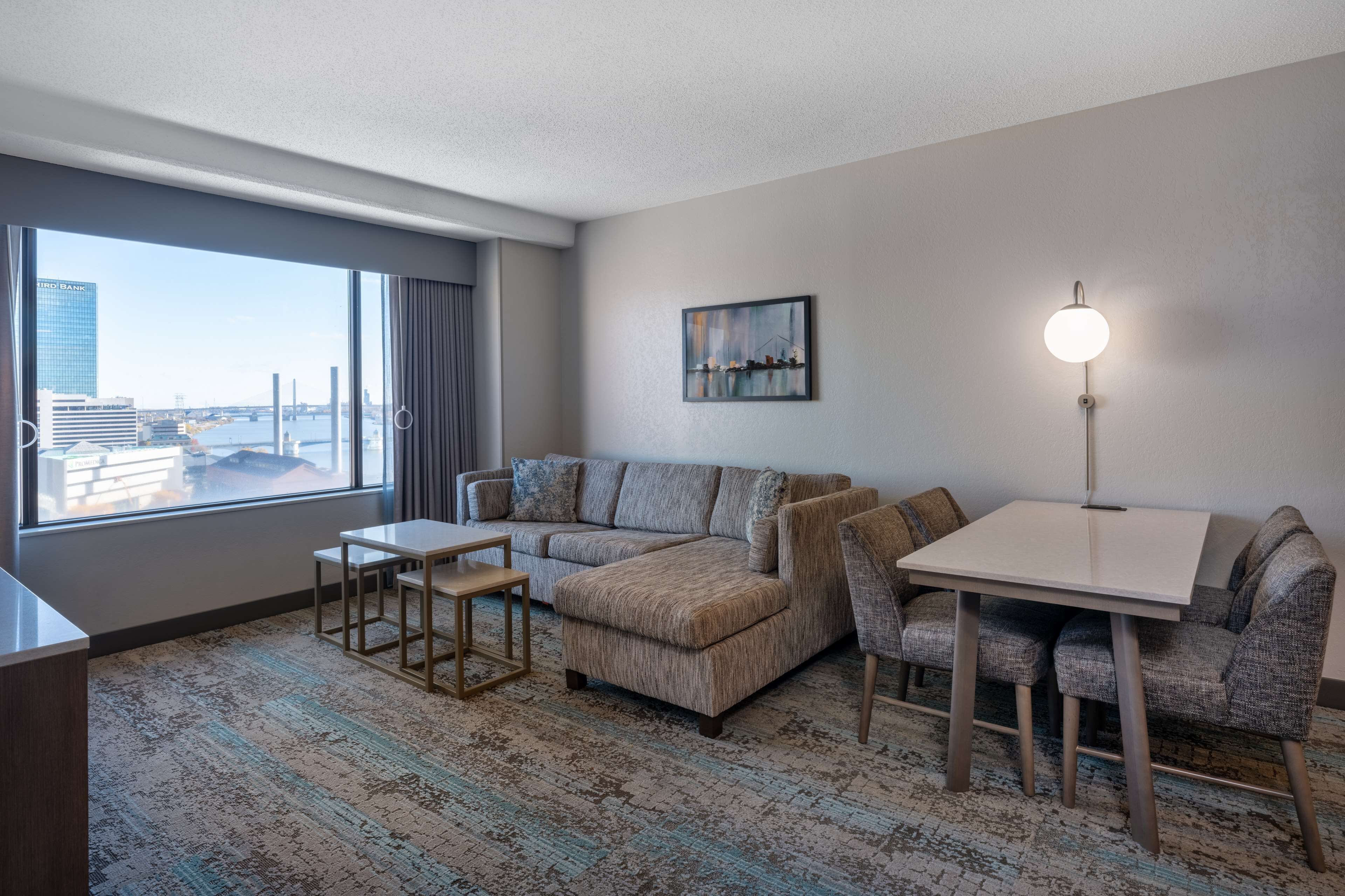 A modern hotel room with a large window, a sofa, a small table, and a lamp.