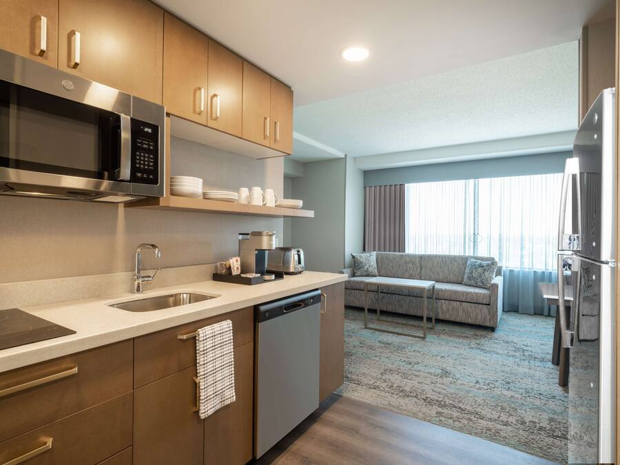 A modern kitchen with stainless steel appliances, a built-in microwave, and a coffee maker. There is a sofa and a dining area in the background.