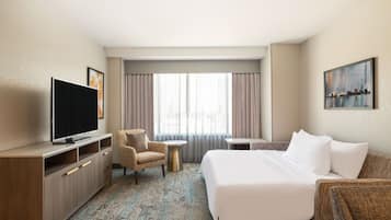 A hotel room with a bed, a TV, a chair, a small table, and a painting on the wall.