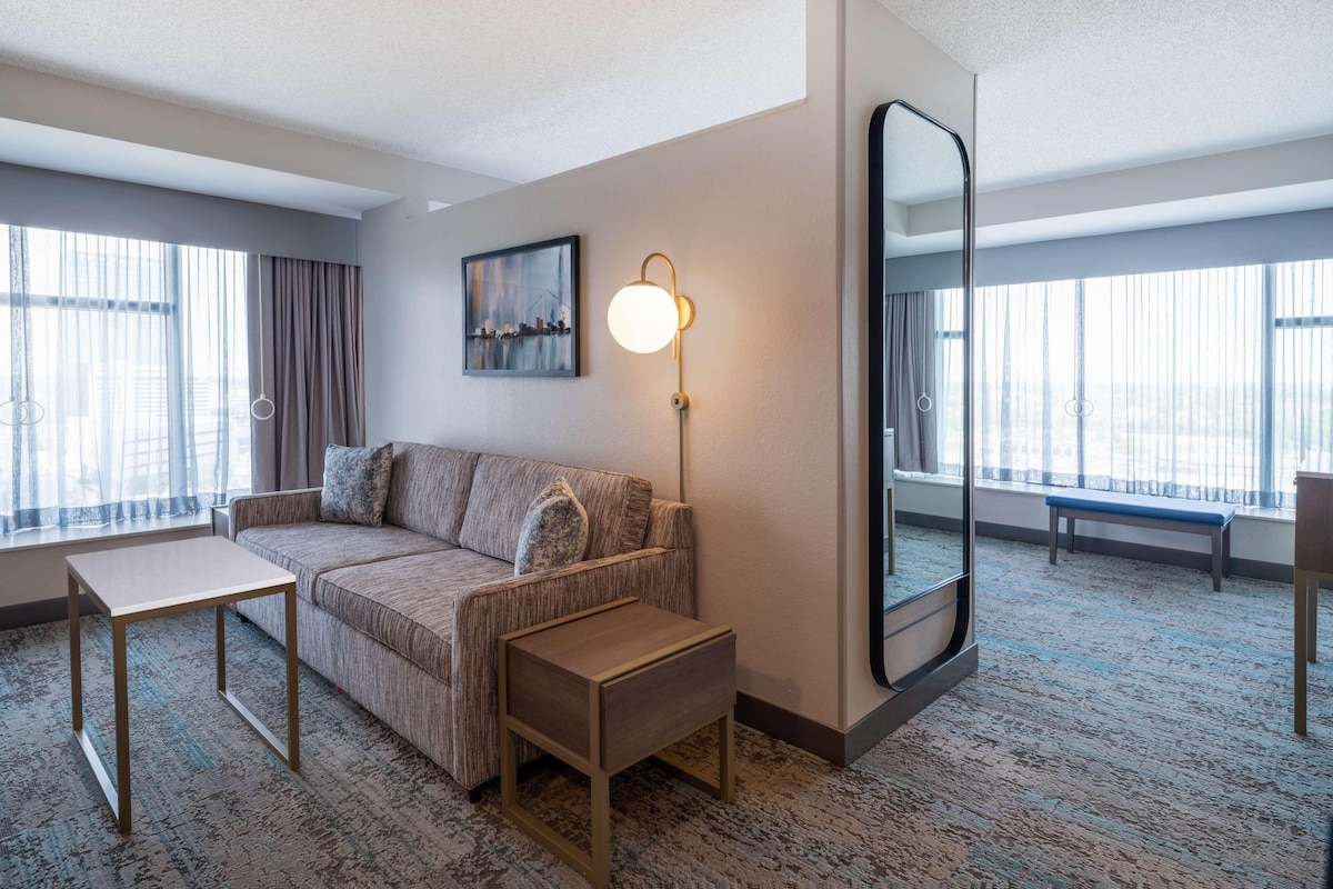 A modern hotel room with a sofa, a small table, a bench, a large mirror, and a painting on the wall.