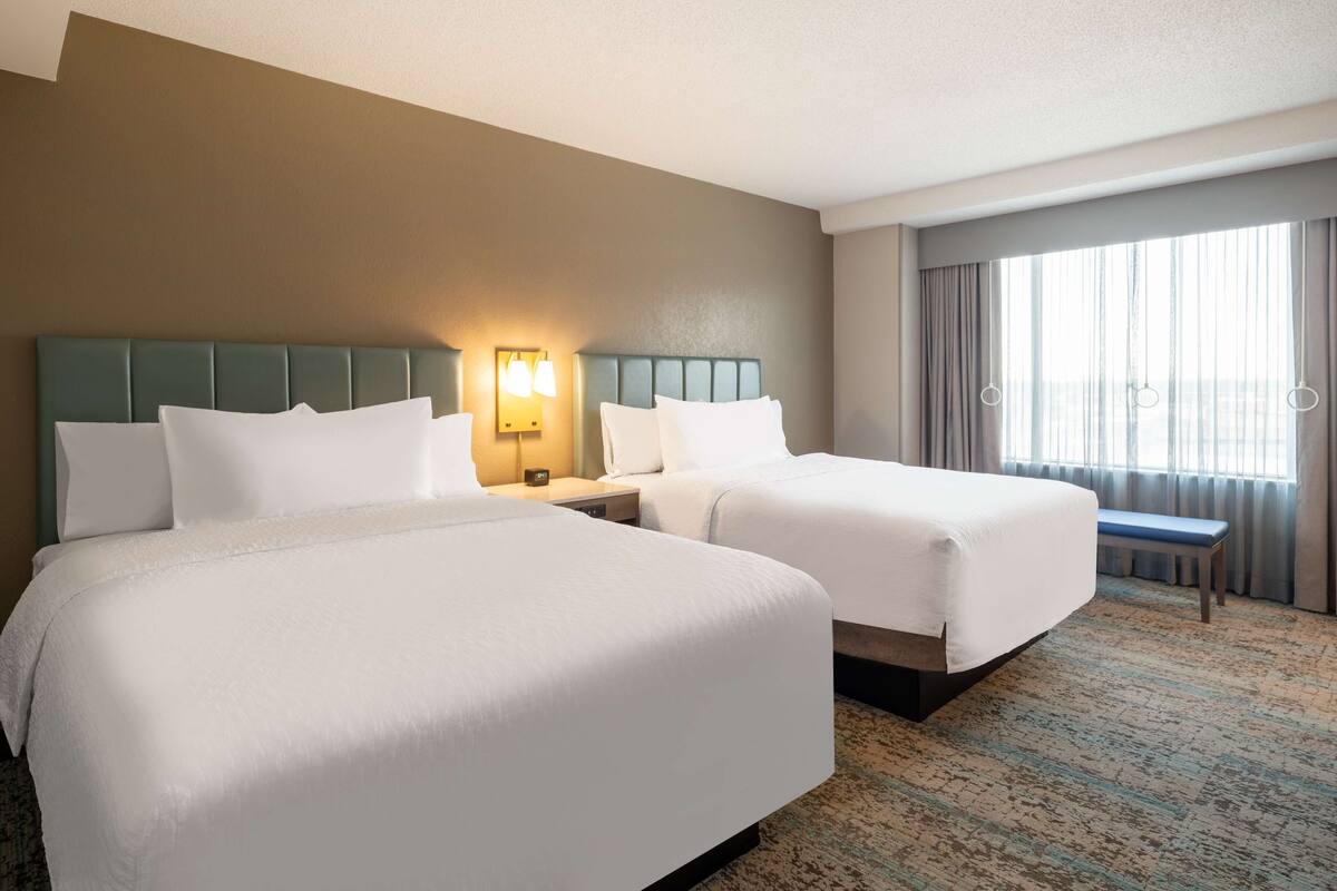 Een hotelkamer met twee bedden, een nachtkastje met een lamp, een bankje en een groot raam met gordijnen.