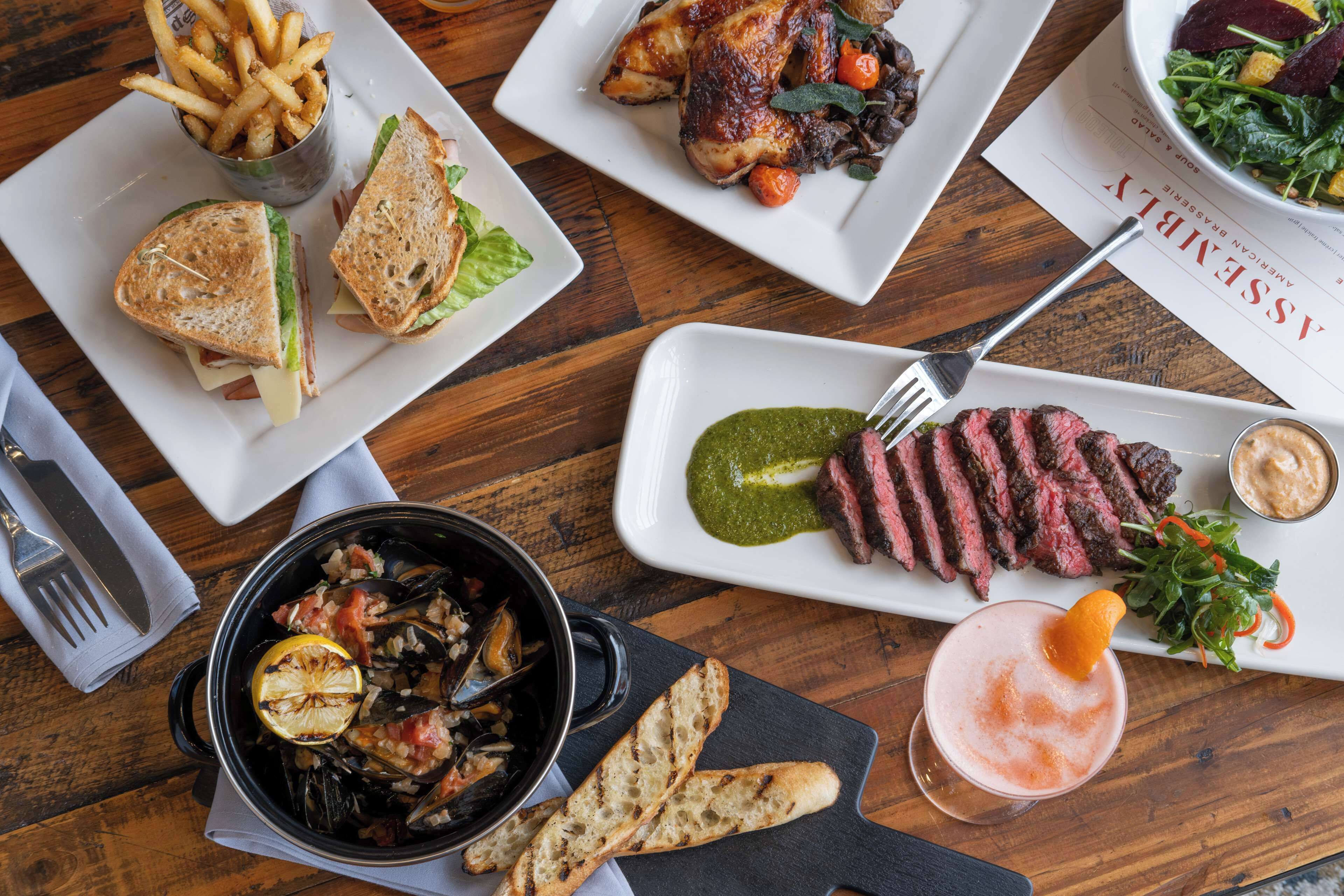 A variety of dishes including a sandwich, steak, and a seafood dish, served on a wooden table.