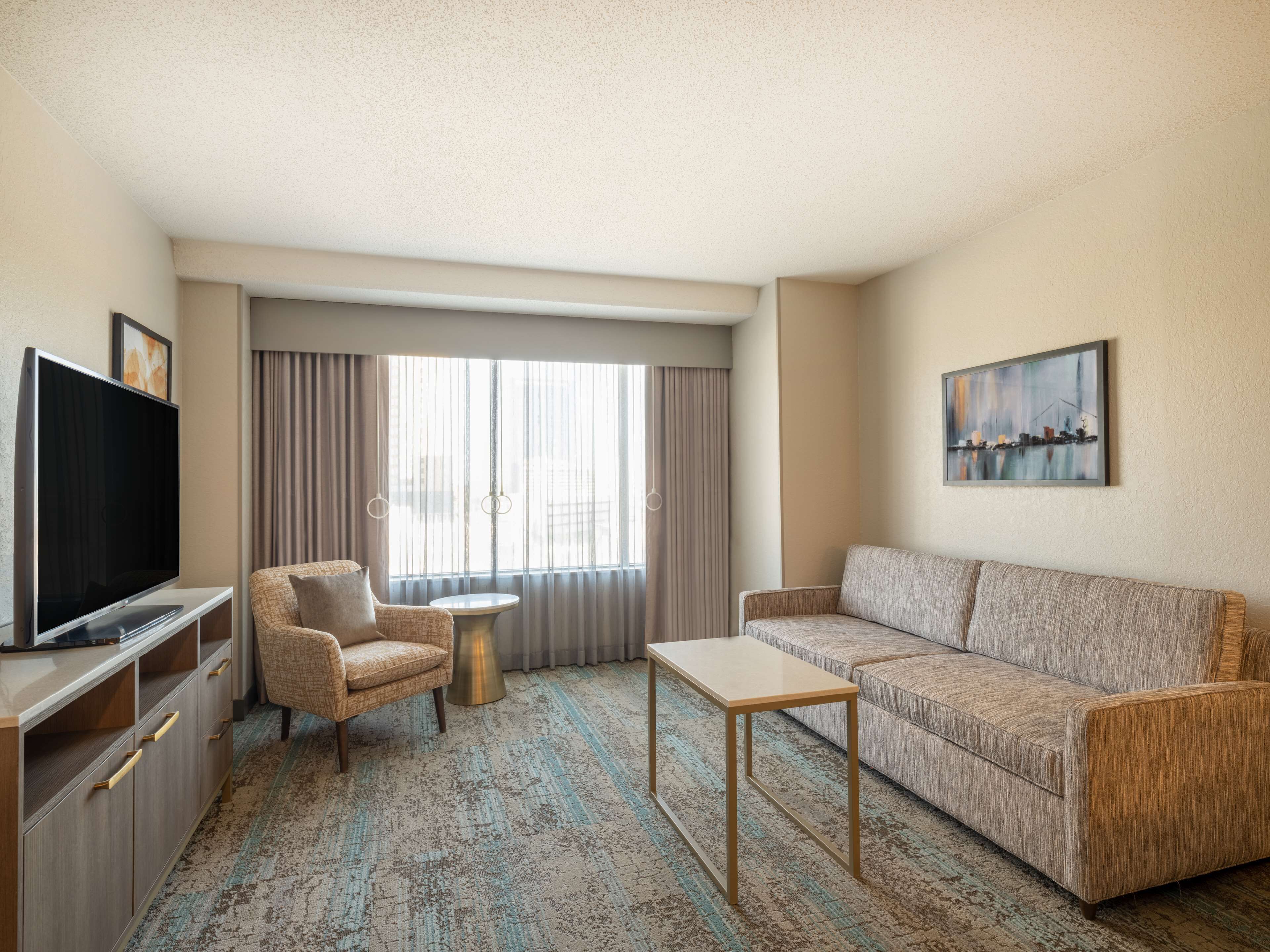 A hotel room with a flat-screen TV, a sofa, a chair, a small round table, and a large window with curtains.