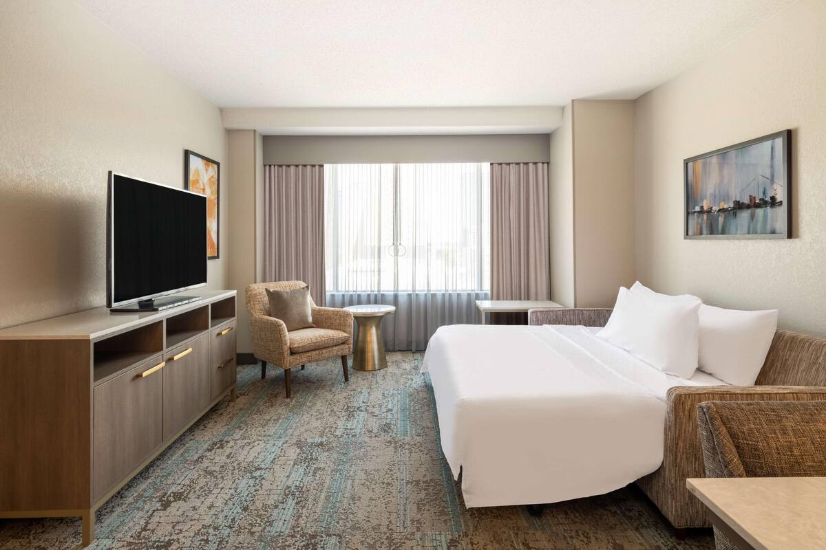 Een hotelkamer met een groot bed, een televisie, een stoel, een tafeltje en een schilderij aan de muur.