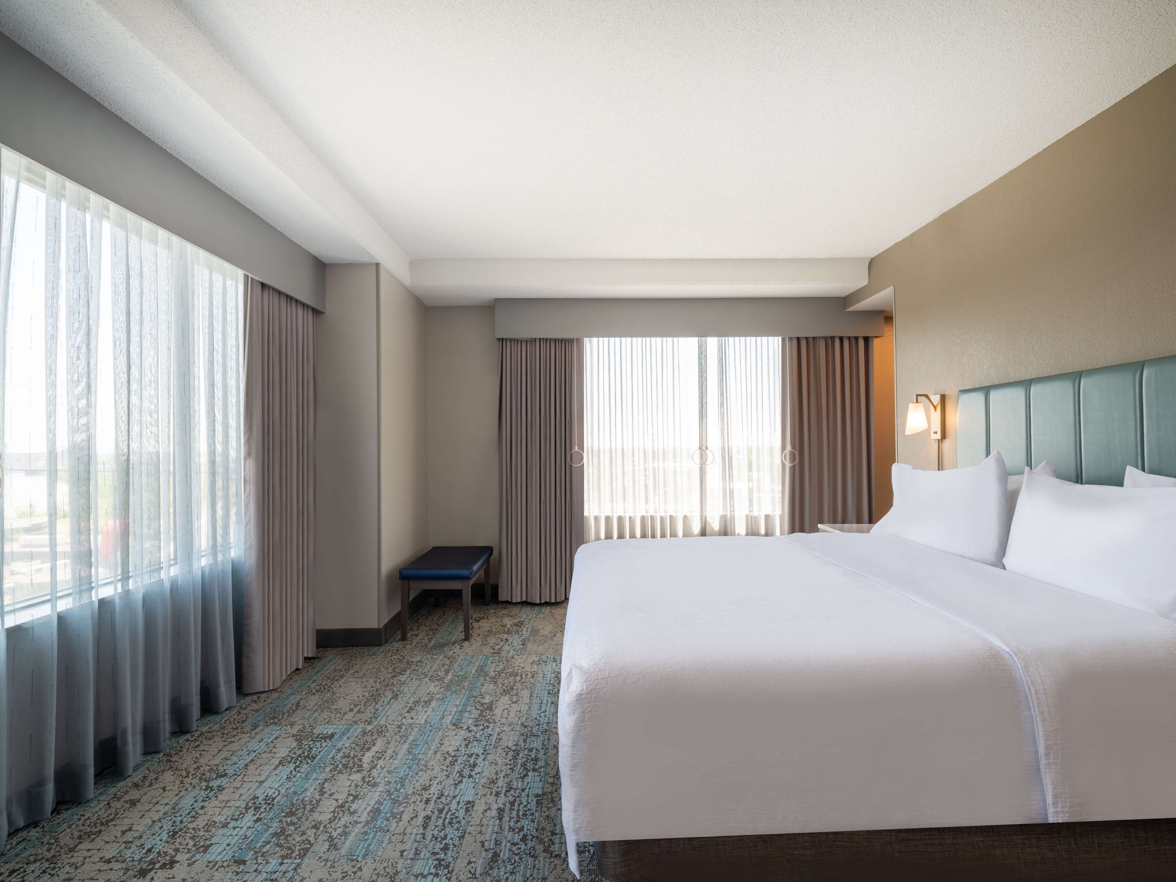 A hotel room with a large bed, a small desk, and a view of the outdoors through large windows.