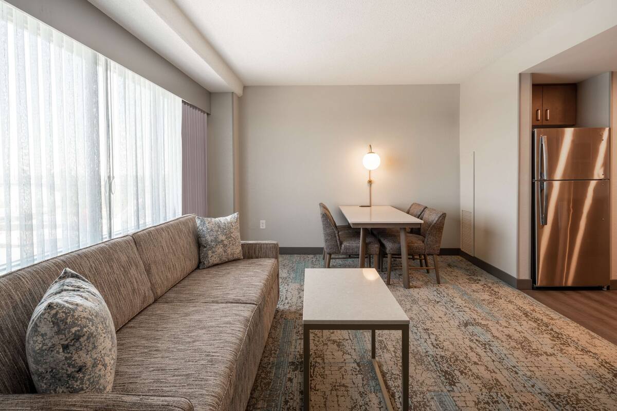 Een moderne woonkamer met een bank, een salontafel en een eethoek met stoelen. Er staat een koelkast en een lamp in de kamer.