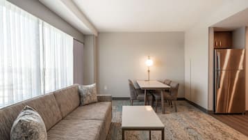 A modern living room with a sofa, a coffee table, and a dining area with chairs. There is a refrigerator and a lamp in the room.
