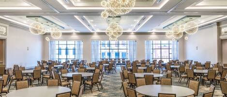 Un amplio salón de conferencias con varias mesas redondas y sillas, ventanas grandes y candelabros de cristal decorativos.