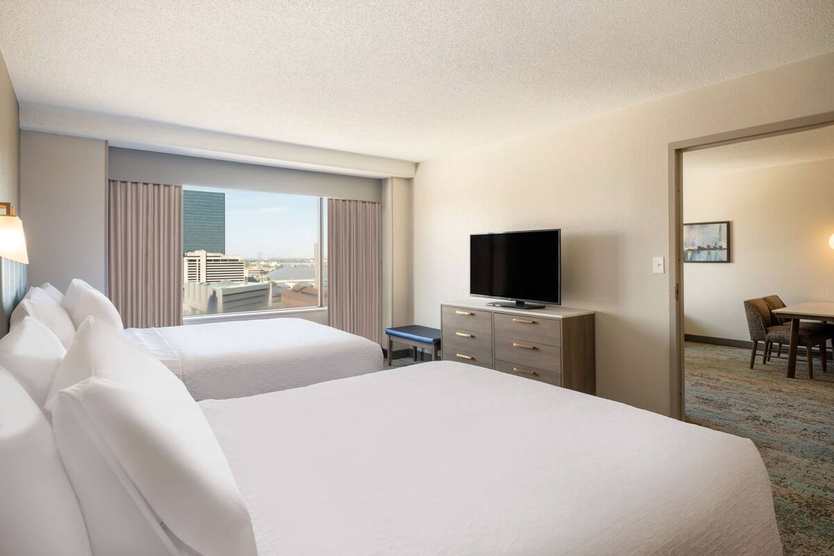 Een hotelkamer met twee bedden, een televisie op een dressoir en uitzicht op gebouwen door het raam.