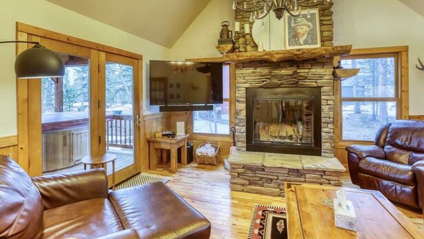 A cozy living room with a stone fireplace, leather sofas, a television, and a deck with large windows.