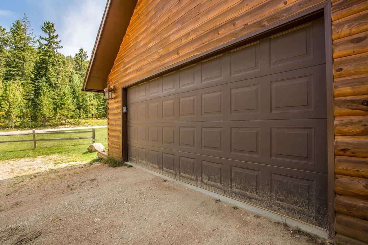 A log cabin with a large garage door, surrounded by a dirt driveway and a wooden fence.