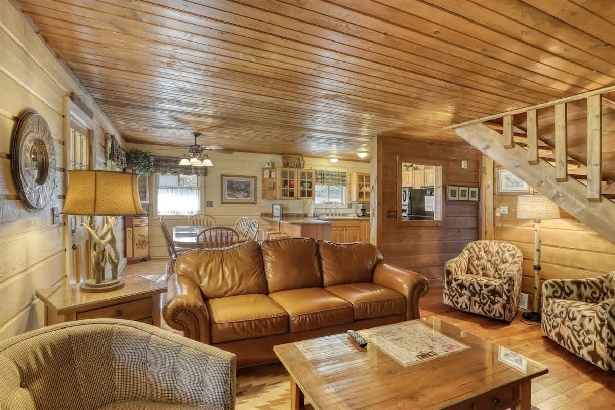 L’intérieur chaleureux d’un chalet avec un plafond en bois, un canapé en cuir, des fauteuils à motifs, un coin repas et un escalier.