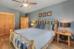 A bedroom with a wooden bed frame, a ceiling fan, a bedside table with a lamp, and a wall with framed pictures.