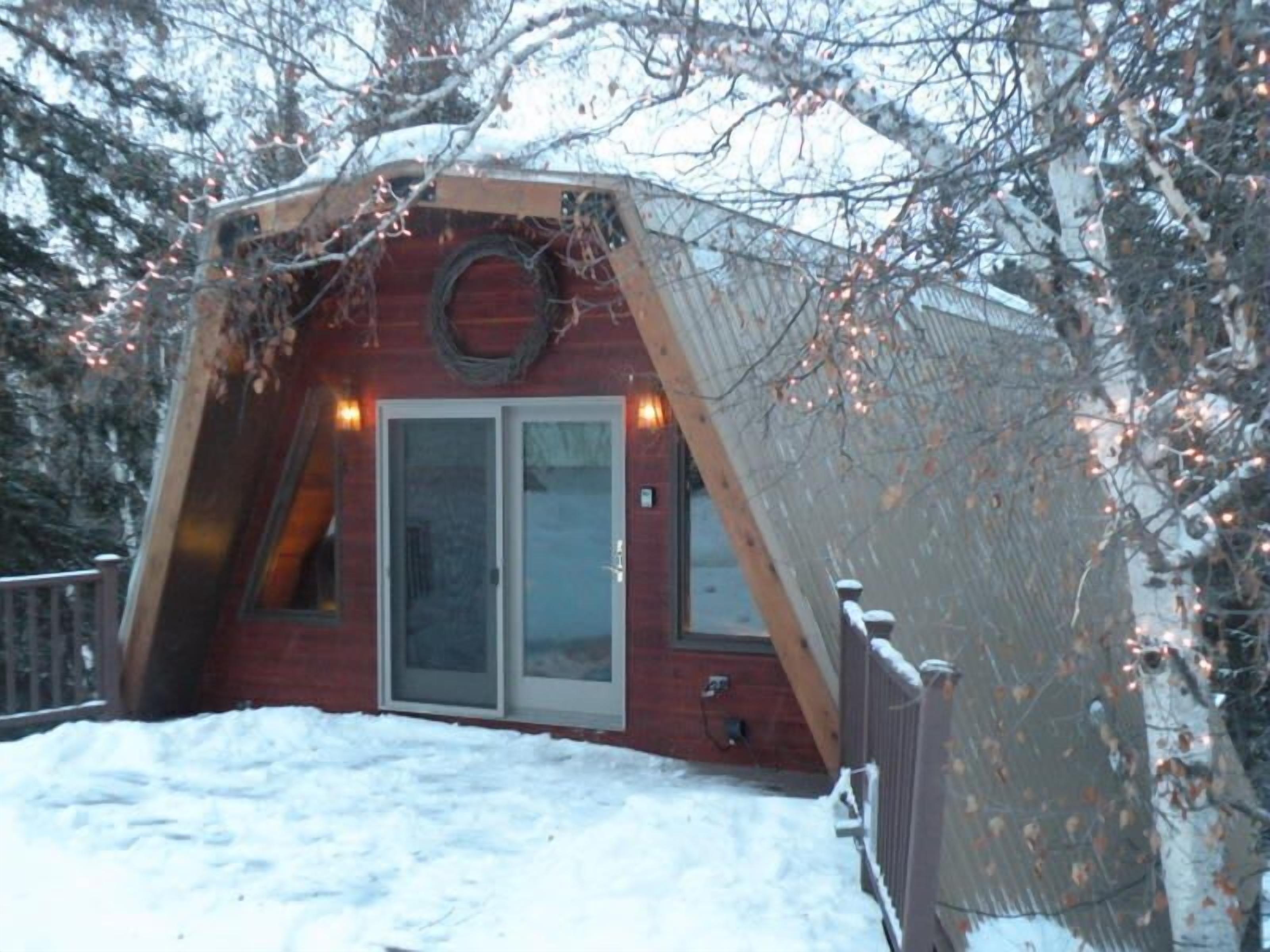 A snow-covered cabin with a unique triangular roof, a glass door entrance, and a wreath on the door.